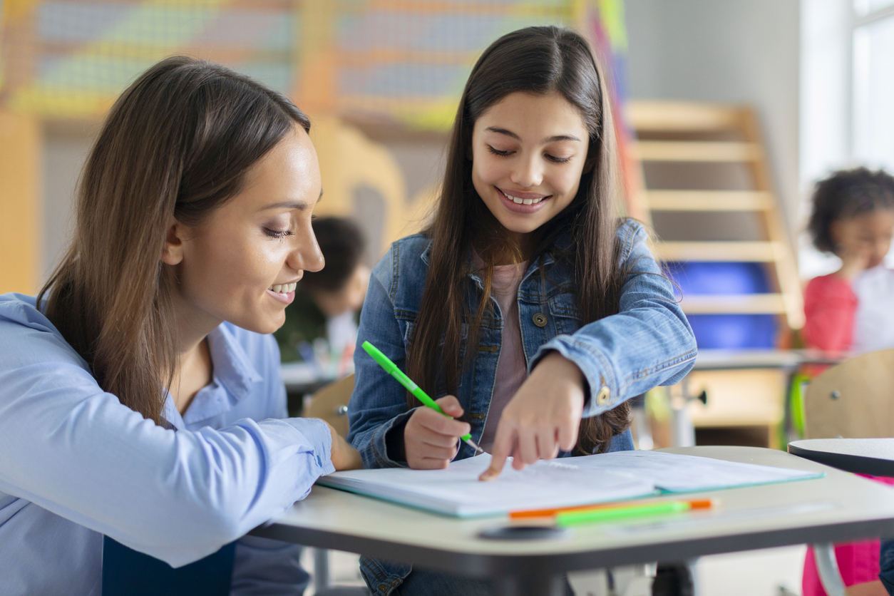 A female teacher kneels down to help a young female student.