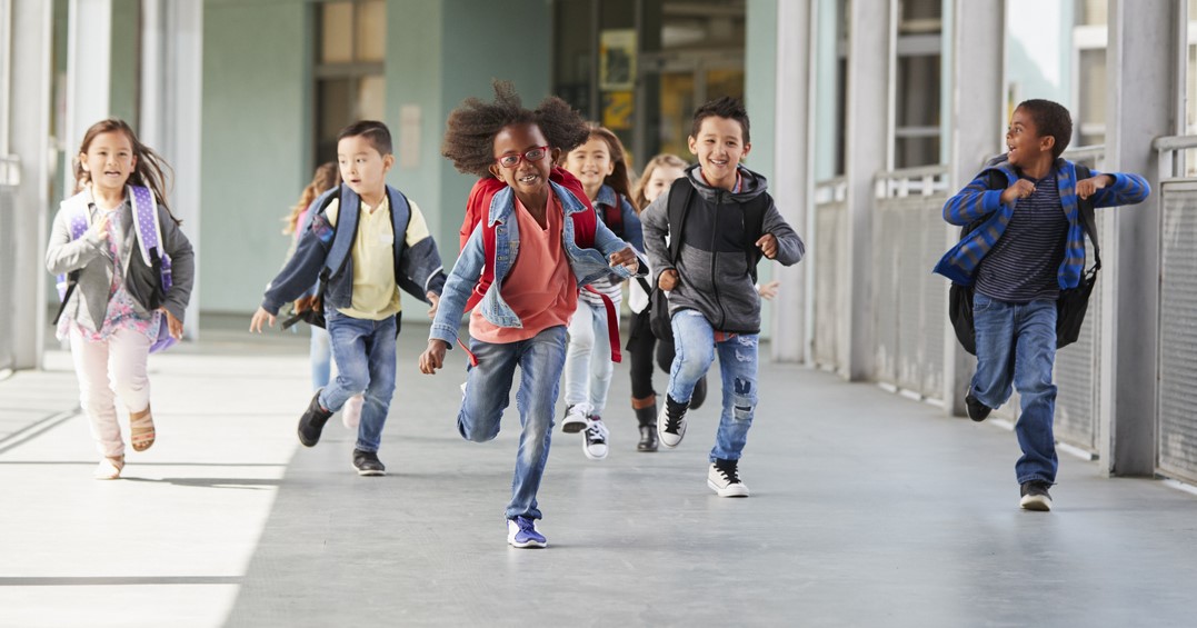Un grupo de estudiantes de primaria con mochilas corriendo por el pasillo de la escuela.