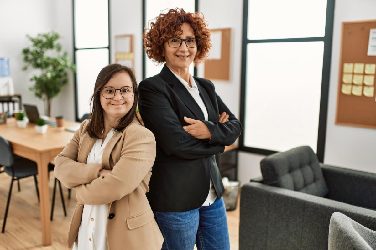 Two women, one with a disability and one without a disability, standing back to back in an office.